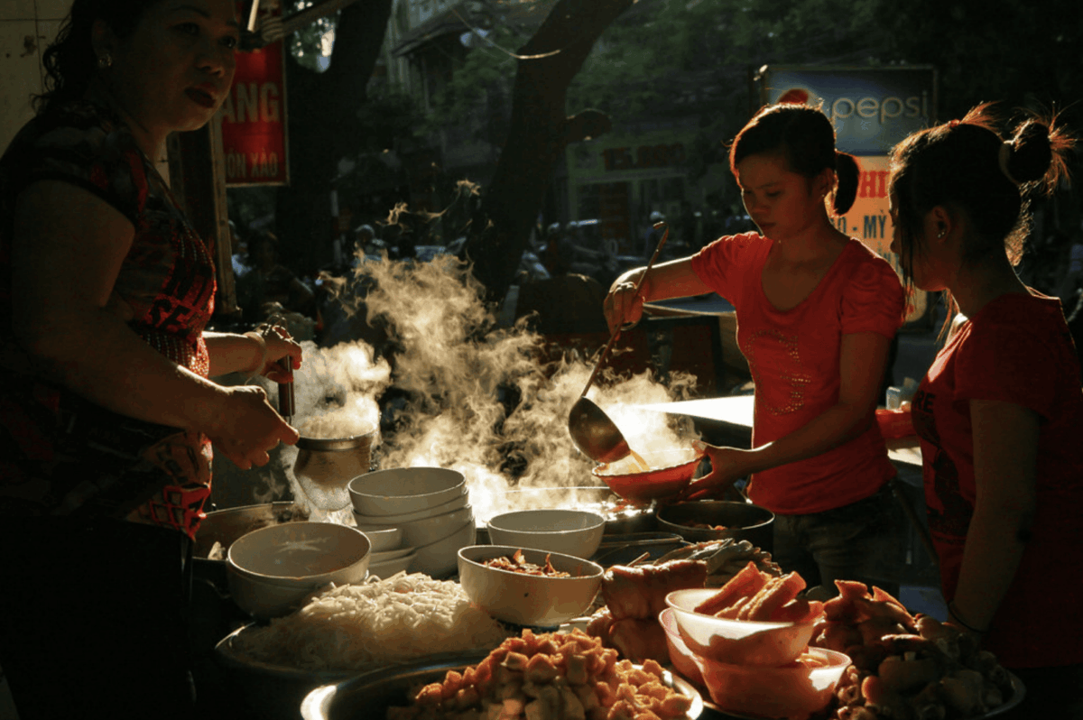 Women serving bowls of Vietnamese street food, with steam rising from the pots in the dim, smoky light.