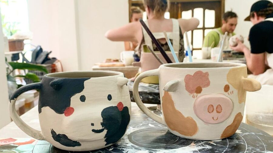 Two handmade ceramic mugs with playful animal designs are displayed at a pottery workshop, with participants working in the background.