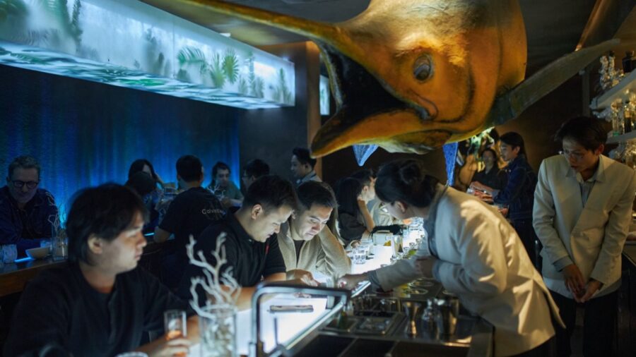 A lively night inside USE Bar Hanoi, where guests sit around the glowing bar counter under dramatic lights and a unique dinosaur-inspired installation.